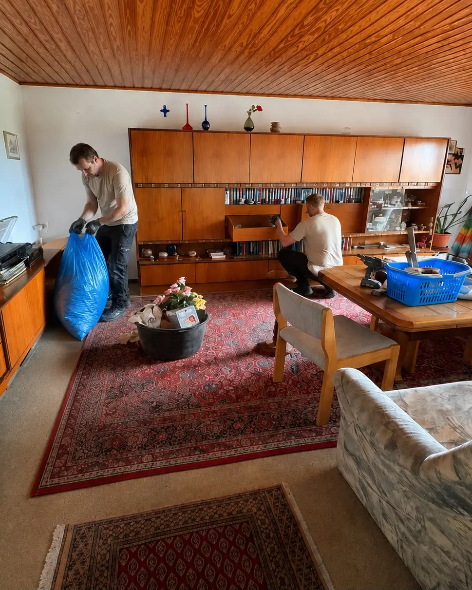 Zwei Mitarbeiter von Kaiserberg Entrümpelung räumen ein Wohnzimmer aus: einer hält einen Müllsack, der andere leert eine Schrankwand aus.