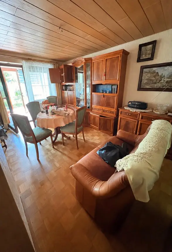 Möbliertes Wohnzimmer mit Holzdecke vor der Entrümpelung: Schrankwand, Esstisch mit Stühlen und Ledersofa.